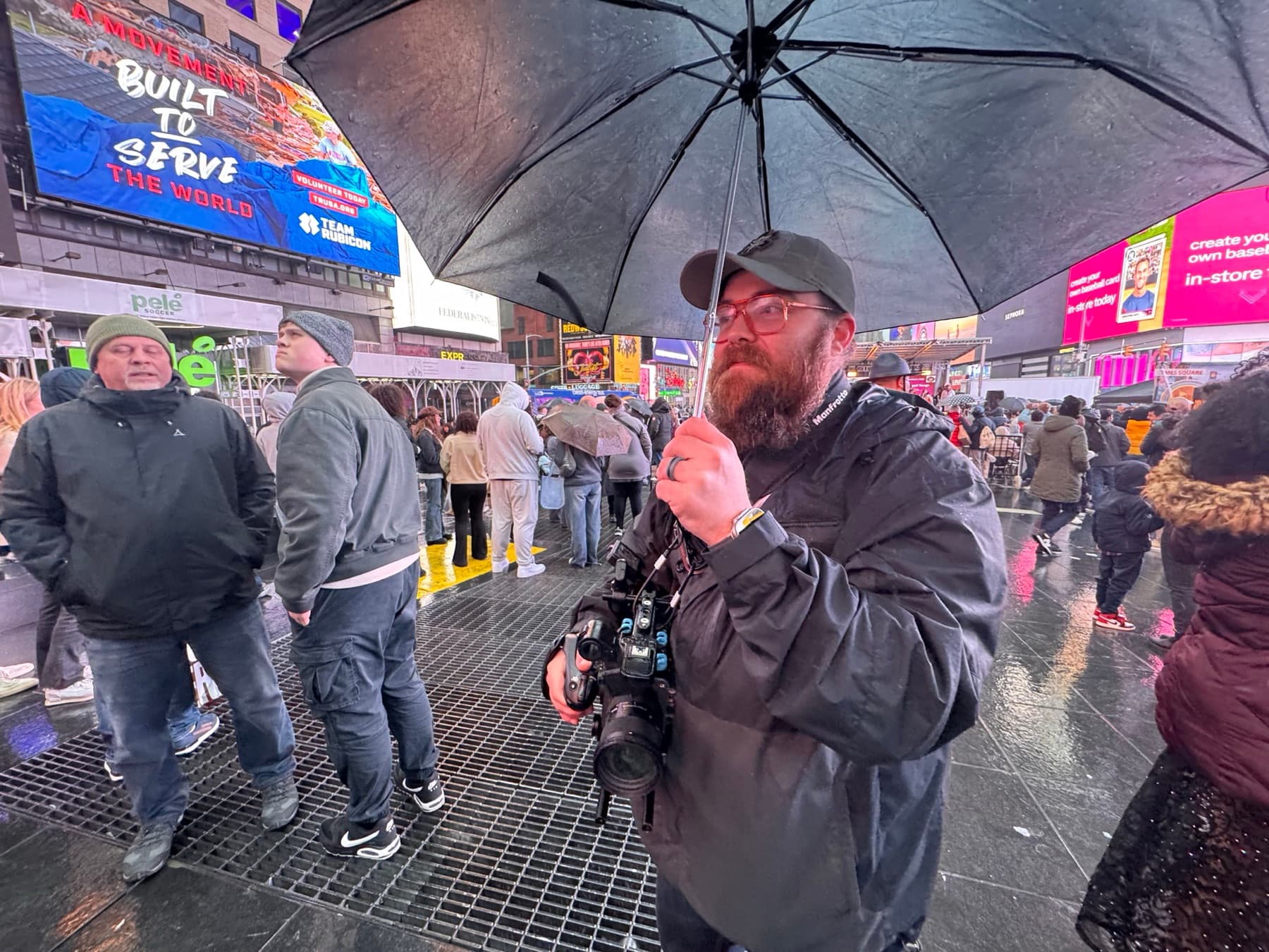 A documentary filmmaker holding a camera beneath an umbrella in rainy Times Square