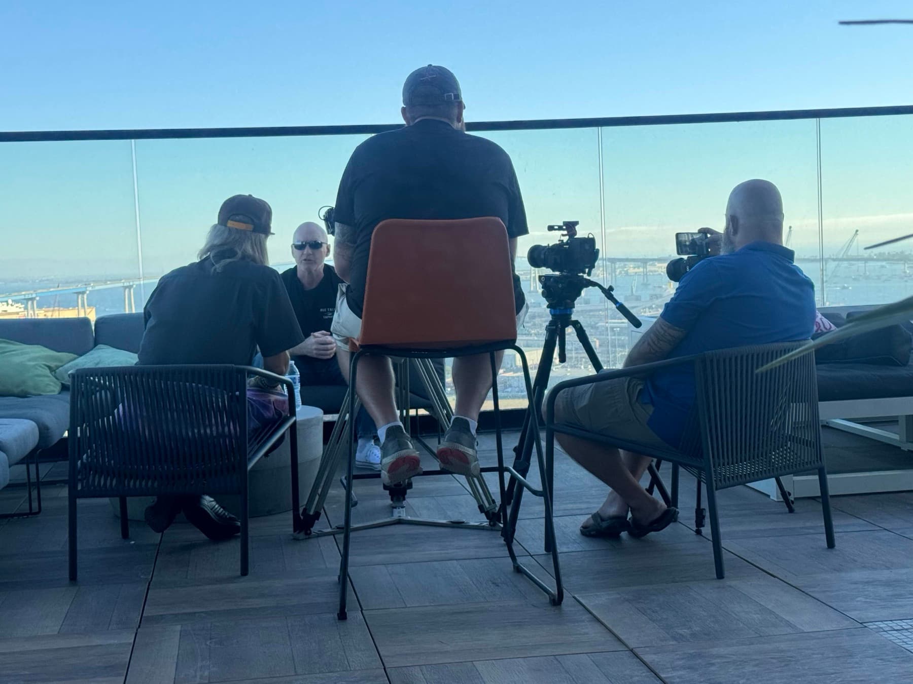 A documentary crew filming an interview with two cameras in a room overlooking a harbor